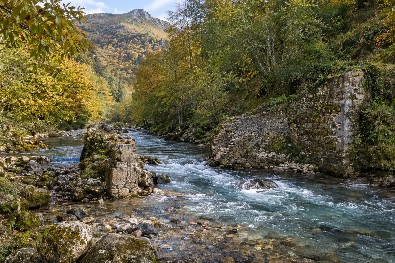 A clear mountain river runs fast over cobbles through a gap in an old, partly demolished stone weir, with moss-covered remains of the wall on one bank and autumn woods on steep slopes behind.