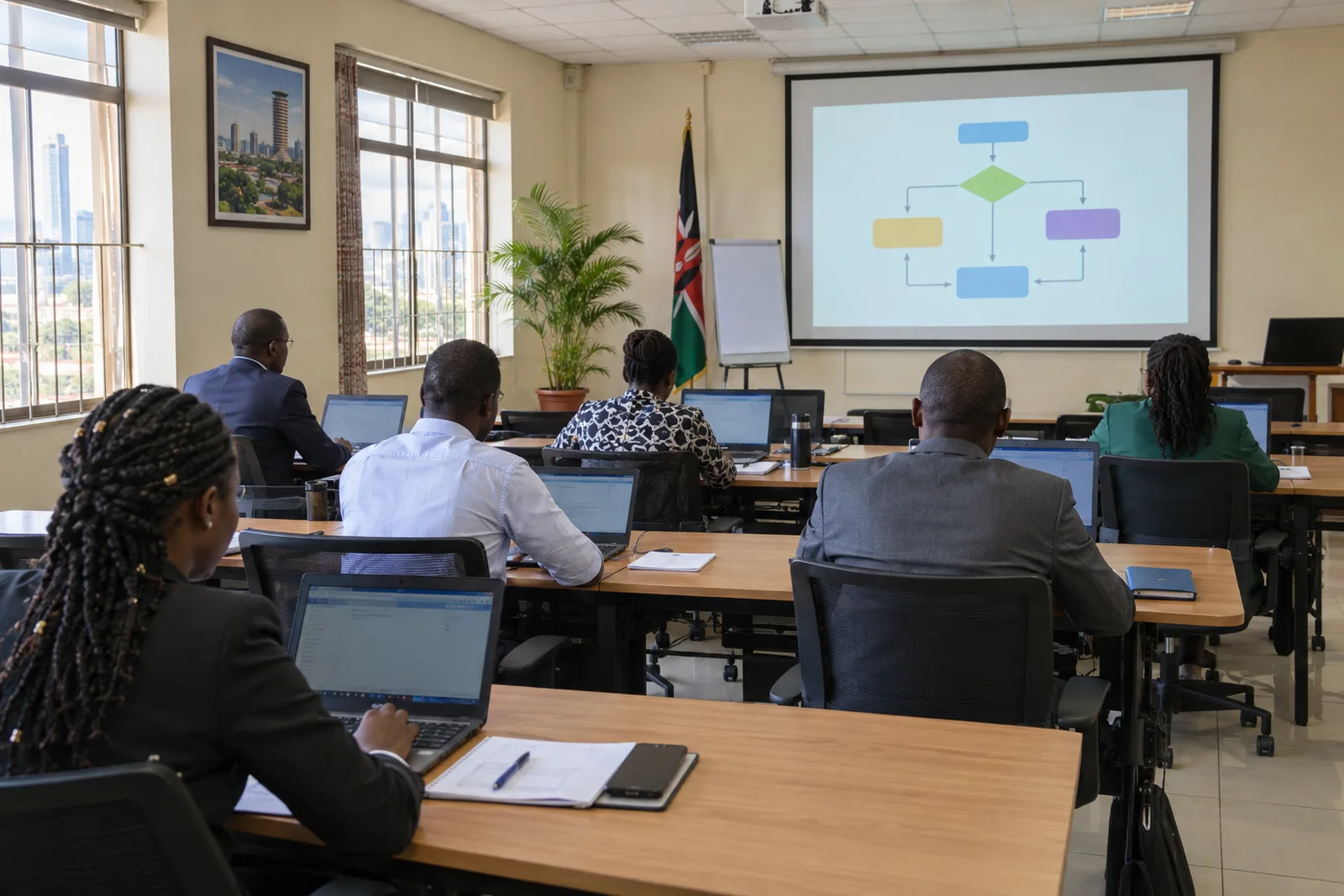 Kenyan civil servants in office clothes, seen from behind, work on laptops at long wooden tables in a bright training room, with a projector screen showing a simple flowchart.
