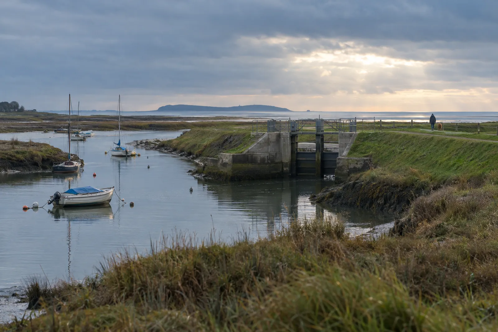 A salt-marsh harbour in early winter, with a grassy flood embankment and a concrete tidal sluice across a creek and small boats on moorings.