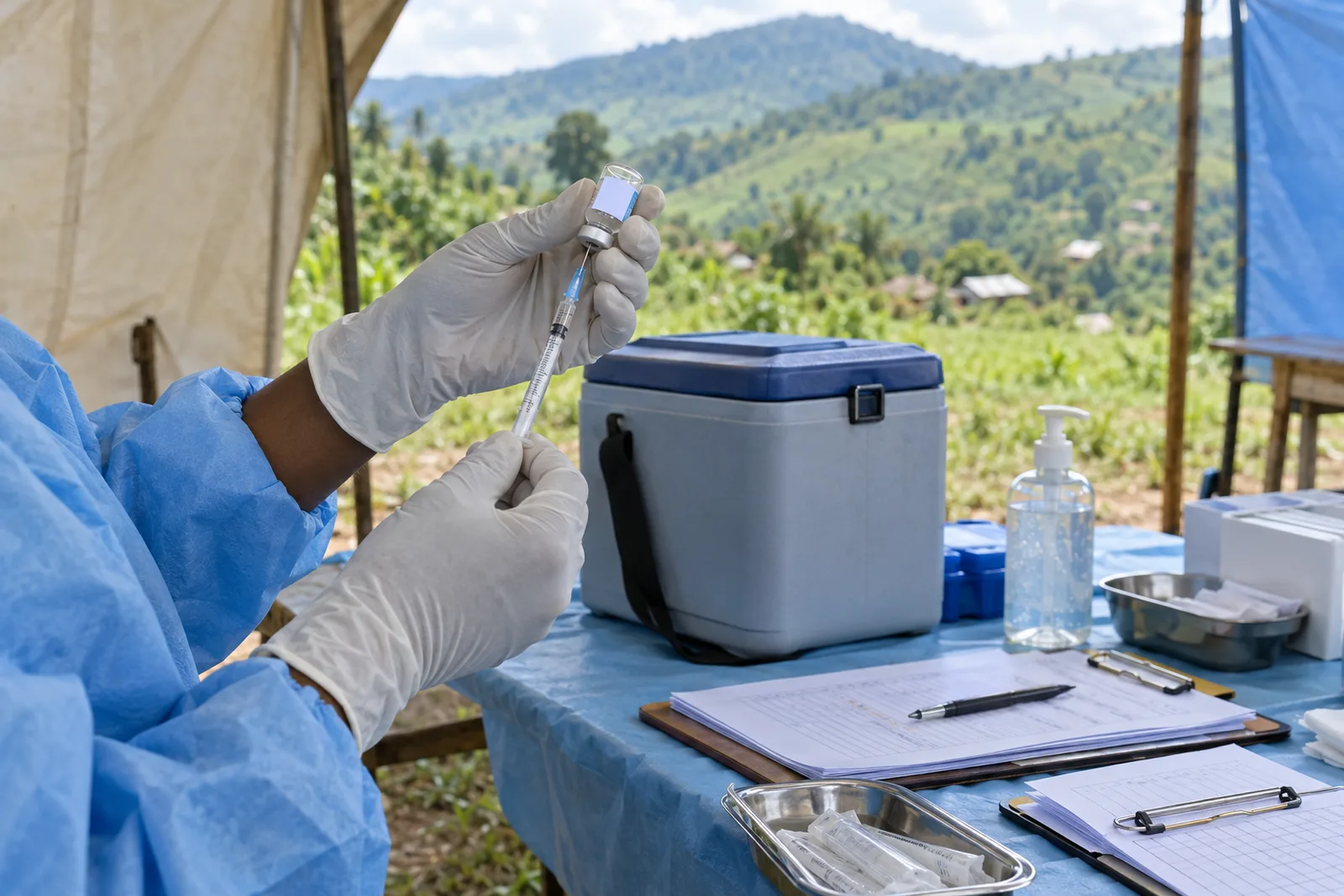 A health worker in a blue protective gown and white gloves draws vaccine from a small vial with a syringe at a table under a tent, beside a cool box, hand sanitiser and clipboards, with green hills behind.